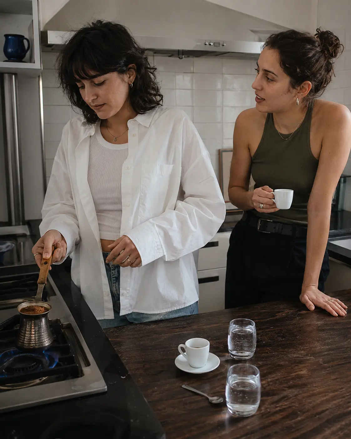 Two people in a kitchen, one heating coffee in a small long-handled pot on a gas hob.